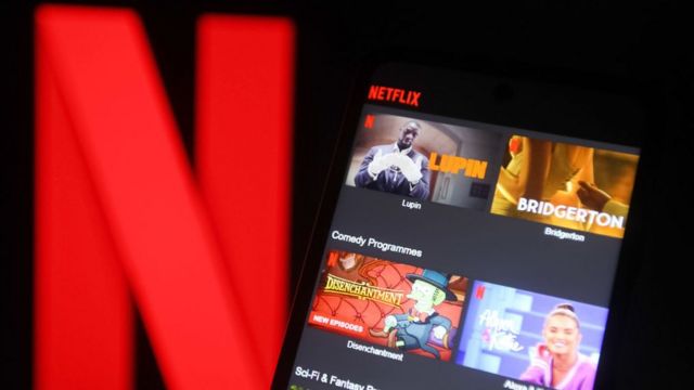 Celular exibindo a tela inicial da Netflix com as séries Lupin e Bridgerton, em frente a um grande logotipo vermelho da marca