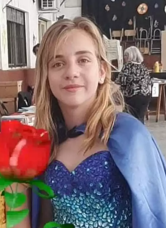 Jovem de cabelo loiro e vestido azul com brilhos, segurando uma rosa vermelha em ambiente interno com mesas ao fundo