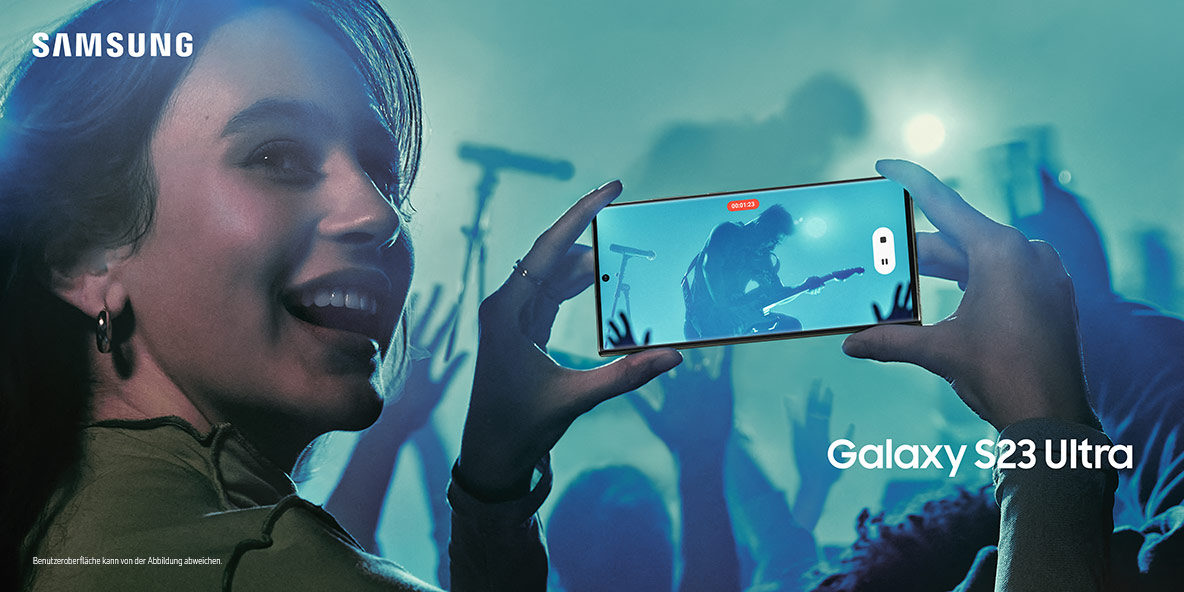 Mulher sorridente filmando show com o Samsung Galaxy S23 Ultra na horizontal, com a tela mostrando um guitarrista no palco