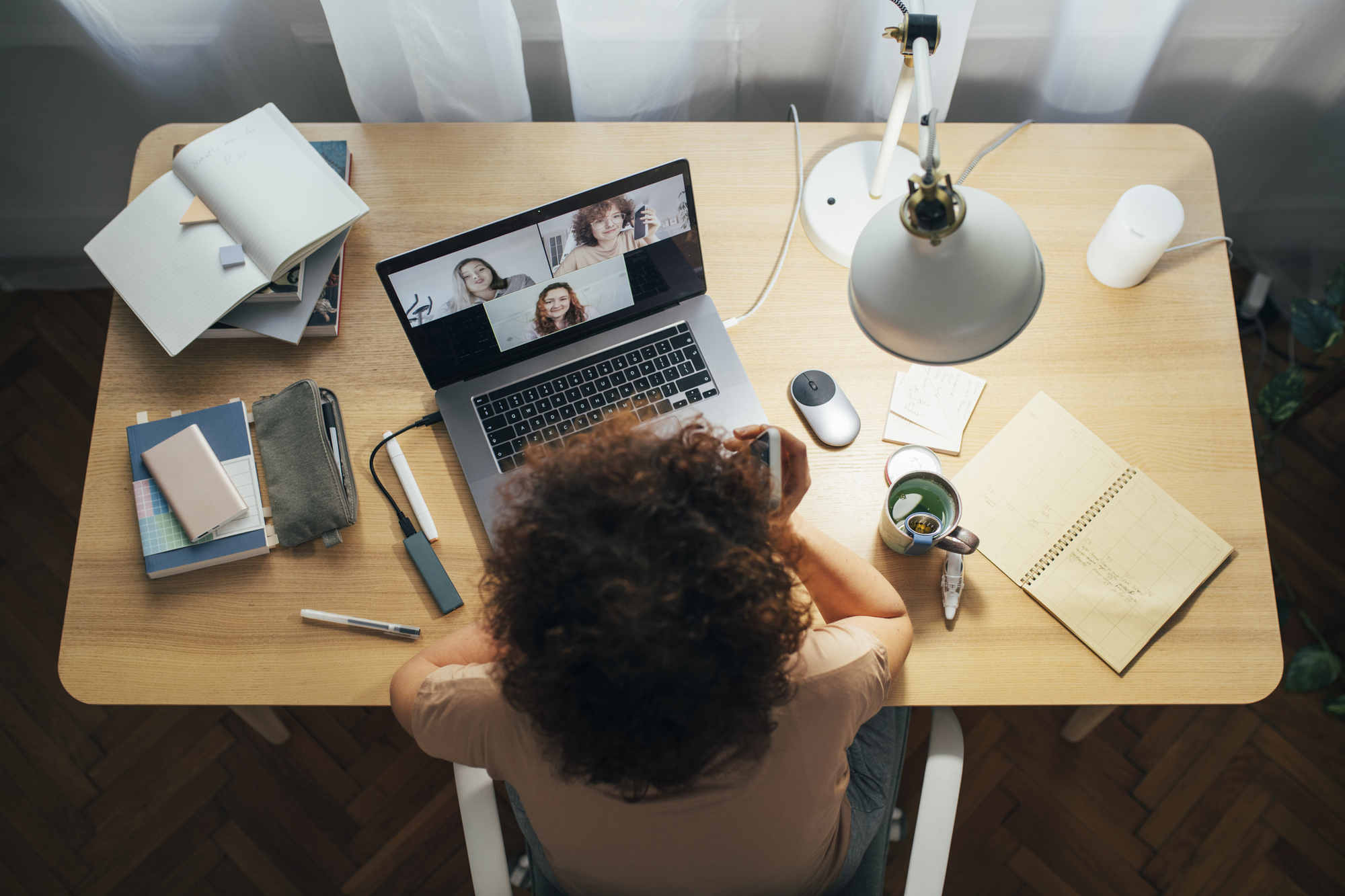 Pessoa de costas em mesa de home office, participando de videochamada em notebook com três colegas na tela, ao lado de cadernos, caneca e mouse