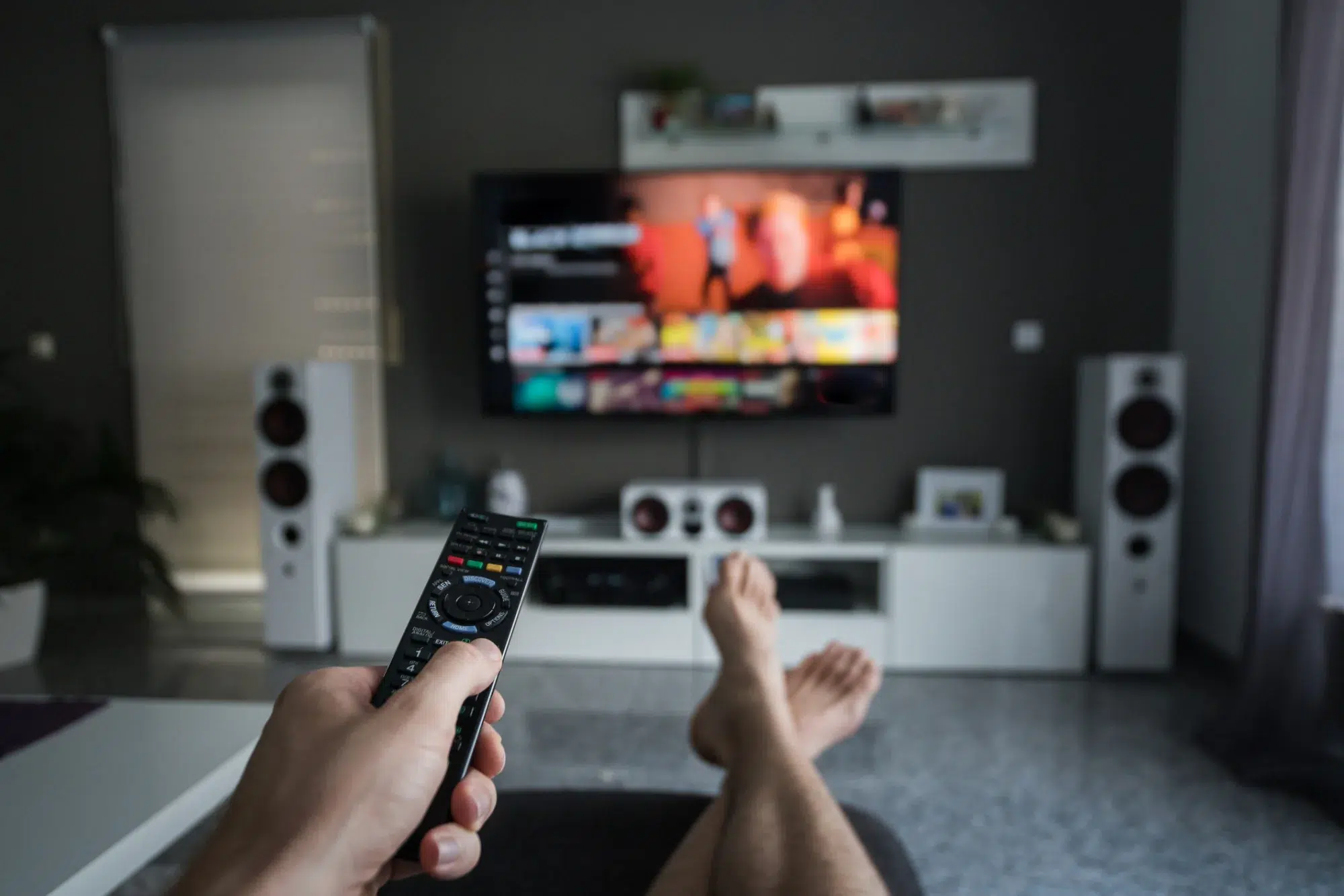 Pessoa deitada no sofá segurando controle remoto e assistindo televisão em uma sala de estar moderna com caixas de som brancas