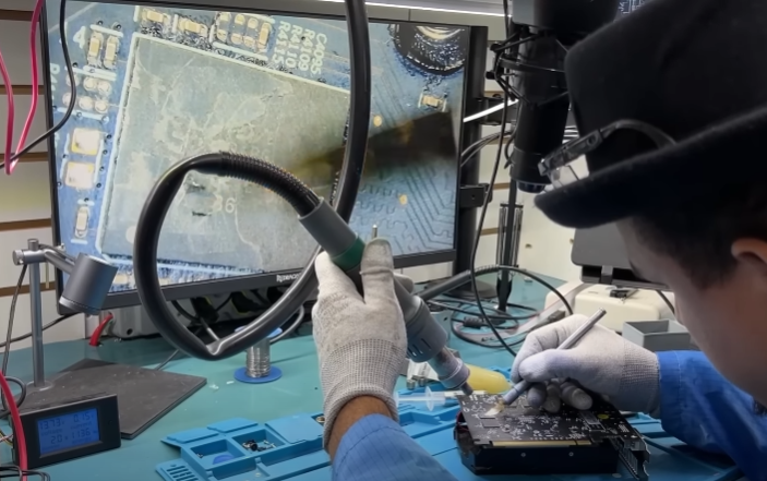 Técnico com luvas e ferramenta de solda reparando uma placa de circuito sobre bancada, com monitor mostrando close da placa ampliada
