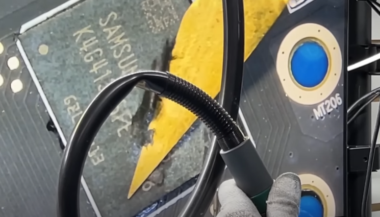 Placa de circuito com chip de memória Samsung sendo aquecido por uma ferramenta de retrabalho, com luva de trabalho e estação de solda ao lado, mostrando o pro…