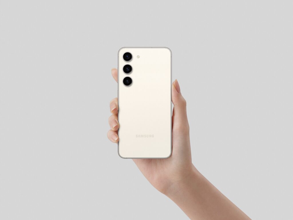 Smartphone Samsung Galaxy S23 na cor bege segurado por uma mão contra fundo cinza, mostrando o traseiro com três câmeras e o logo Samsung
