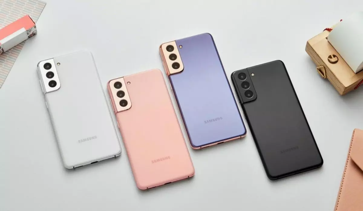 Quatro smartphones Samsung Galaxy S21 enfileirados de costas, nas cores branco, rosa, lilás e preto, sobre uma superfície clara