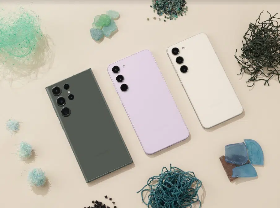 Três smartphones Samsung Galaxy S23 nas cores verde, lilás e creme, vistos de costas sobre fundo bege com fragmentos de vidro e fios reciclados