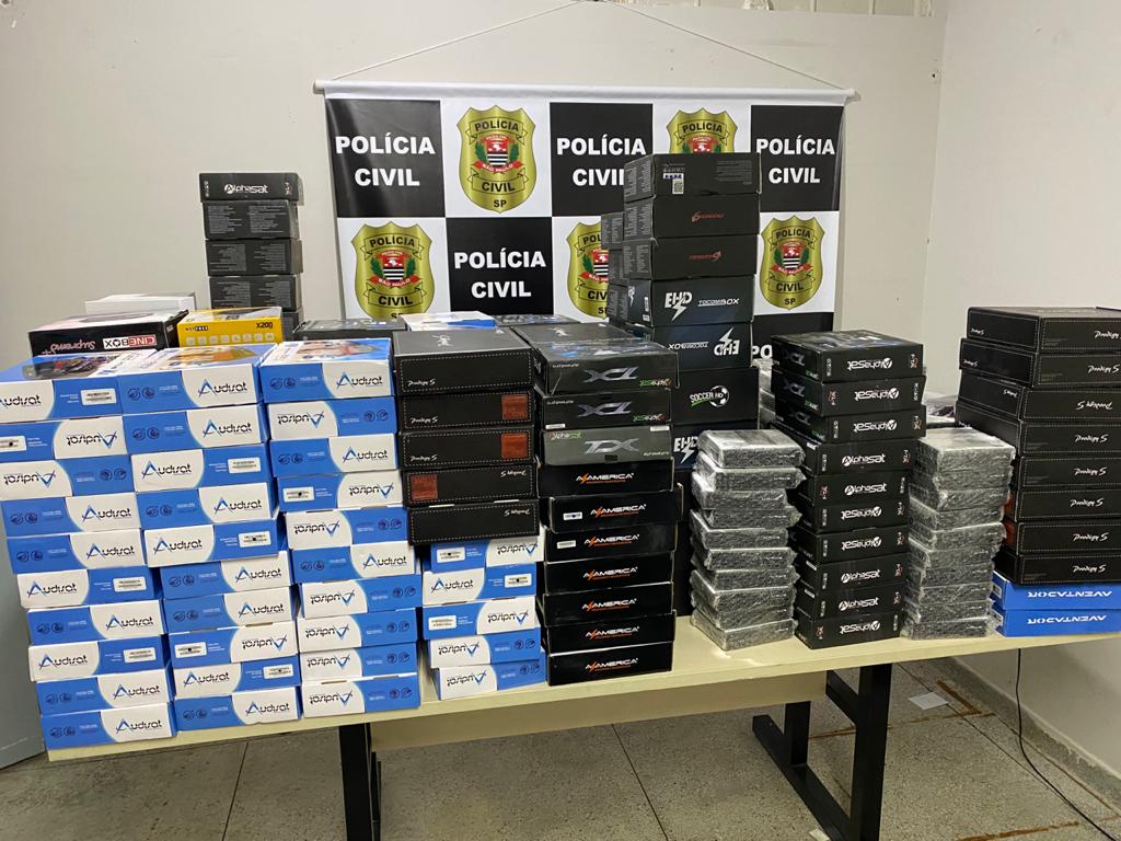 Caixas de TV Box piratas empilhadas sobre mesa diante de banner da Polícia Civil de SP, apreendidas em operação contra IPTV ilegal