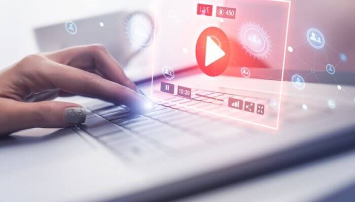 Mão digitando em um notebook com uma interface holográfica de player de vídeo ao vivo, mostrando botão de play, contador de 450 espectadores e ícones de compar…