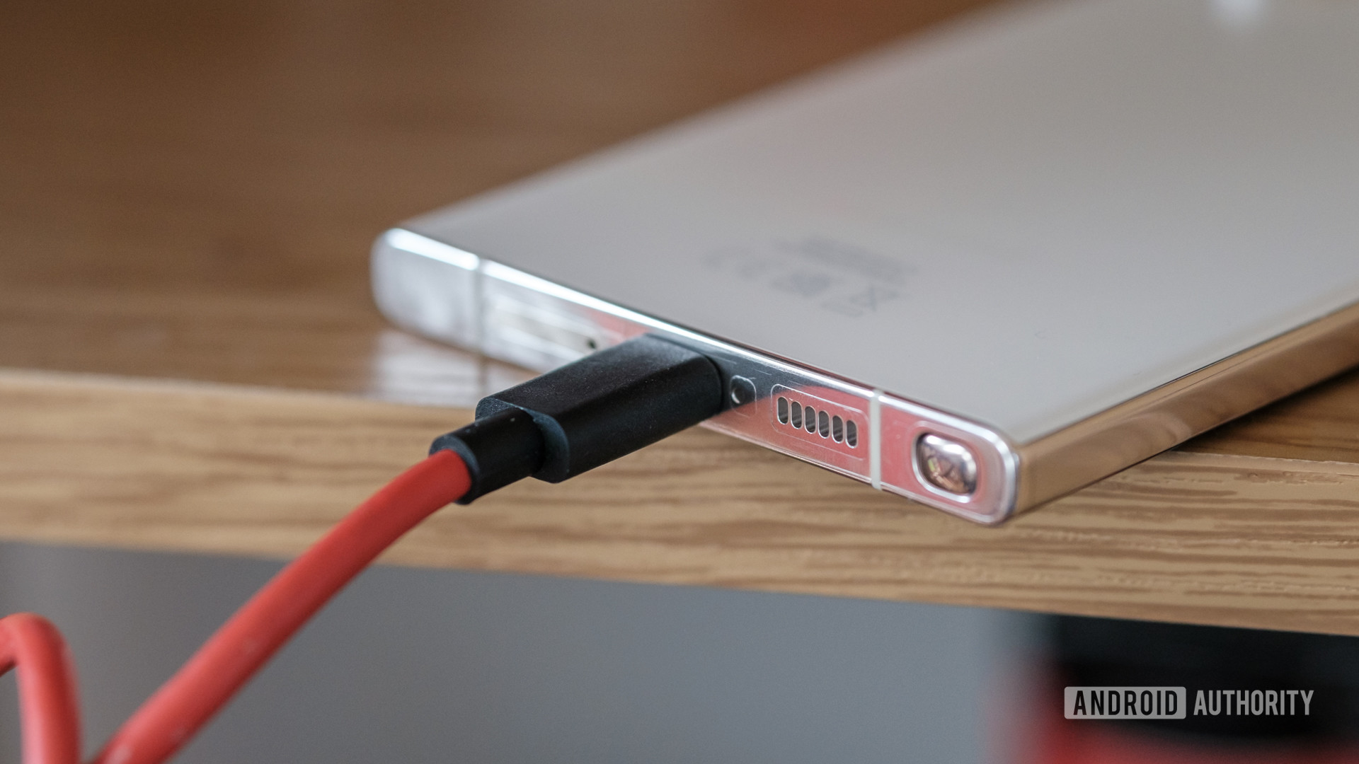 Smartphone prateado com cabo USB-C vermelho conectado à porta inferior, apoiado sobre uma mesa de madeira