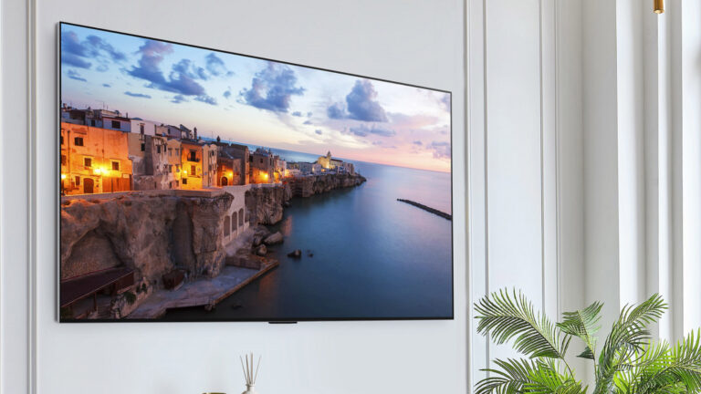 Televisor OLED montado na parede de sala branca, exibindo paisagem de vilarejo costeiro ao entardecer, com planta ao lado