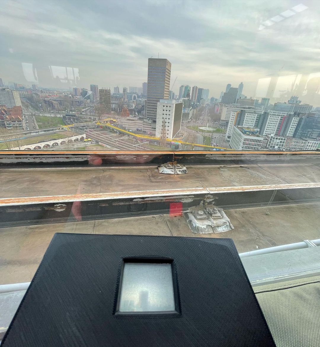Vista de uma cidade pela janela, com um prédio alto ao centro e, em primeiro plano, a câmera preta de uma máquina fotográfica apontada para a paisagem