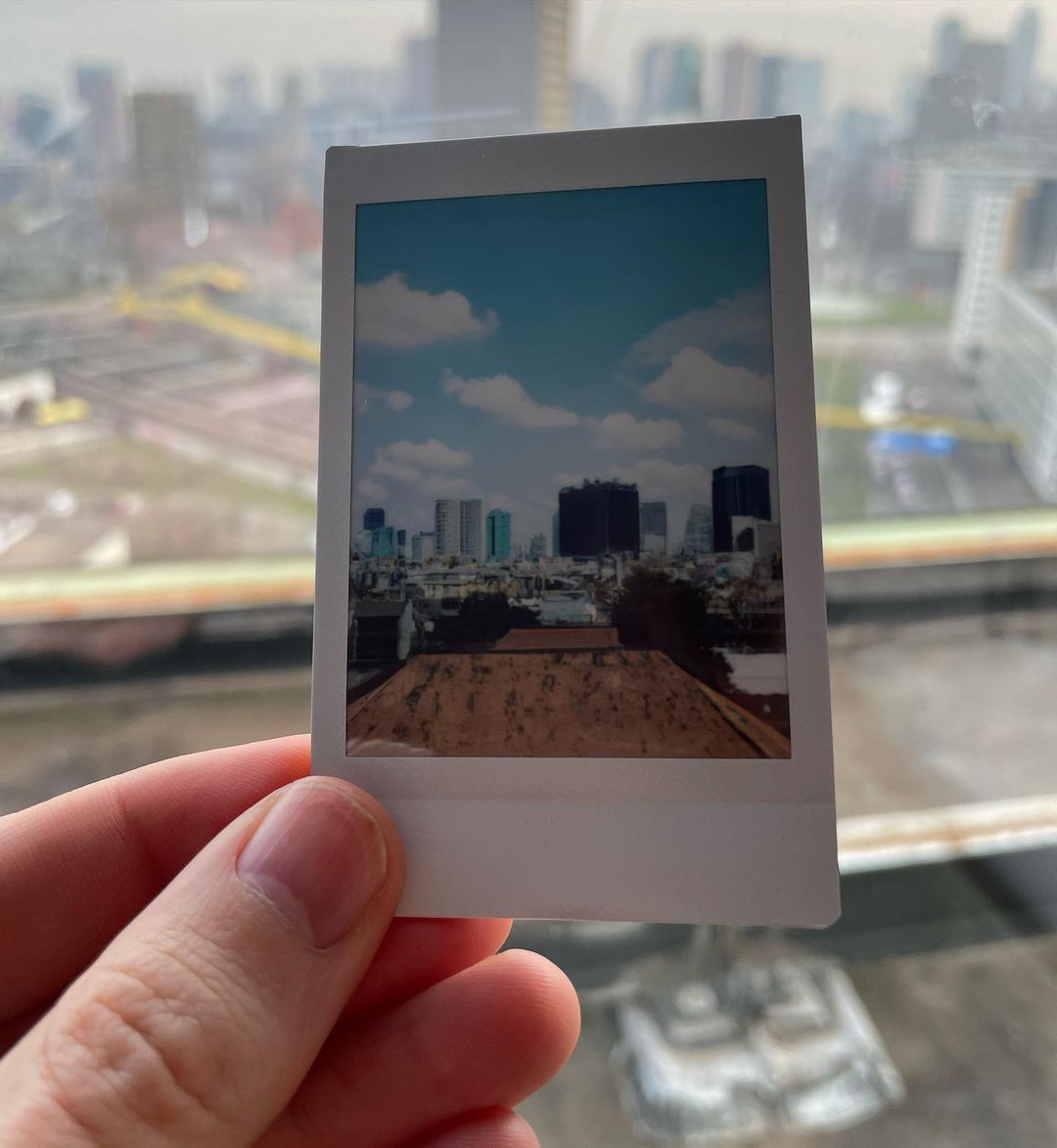 Mão segurando uma foto instantânea com vista de prédios sob céu azul com nuvens, em frente a uma janela com paisagem urbana desfocada ao fundo