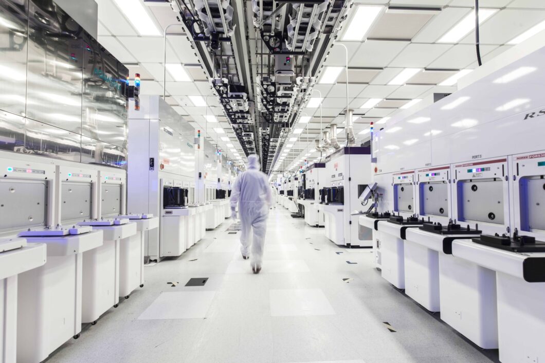 Técnico com macacão branco caminha por corredor de sala limpa de fábrica de semicondutores, ladeado por máquinas brancas de produção de chips