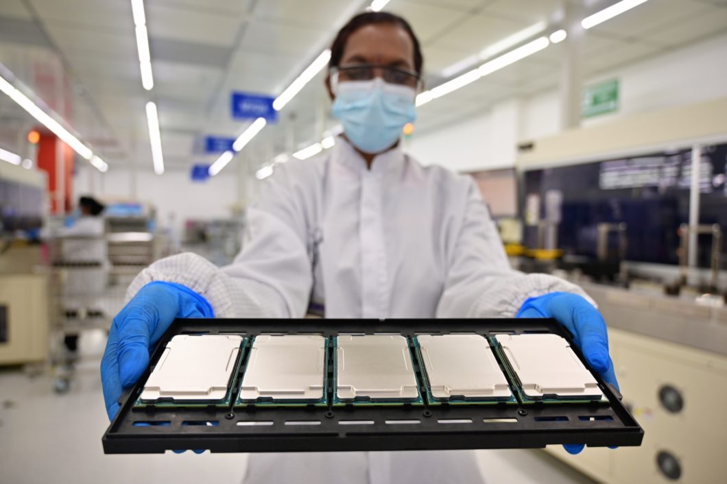 Técnico de uniforme branco e máscara azul segura bandeja preta com processadores Intel Xeon em ambiente de fábrica iluminado