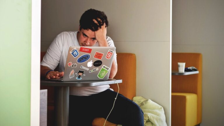 Homem sentado com a mão na cabeça, aparentando preocupação diante de um notebook com adesivos de tecnologia como PHP, Git, Node e GitHub