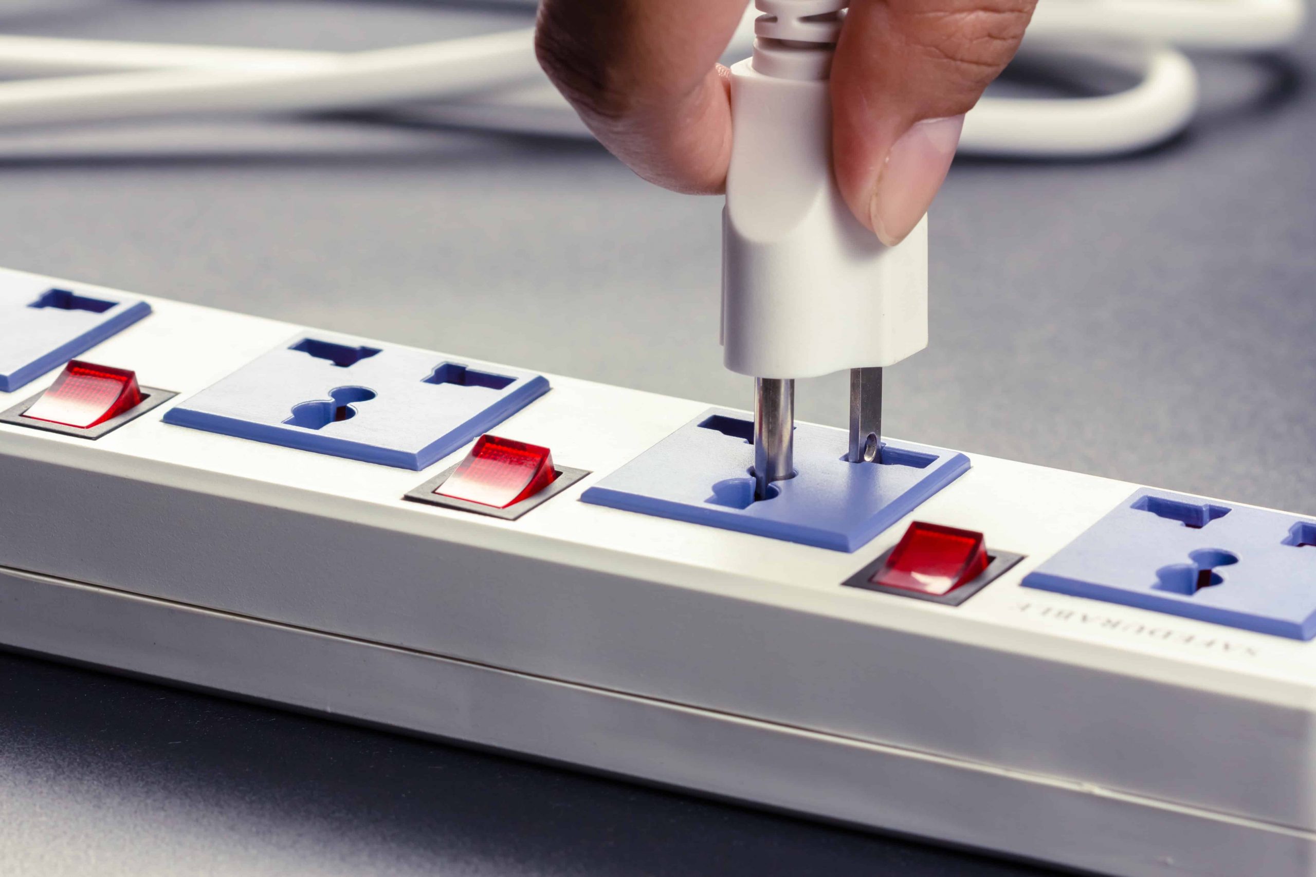 Mão conectando um plugue branco a uma régua de tomadas branca com interruptores vermelhos e tomadas azuis, de modelo universal