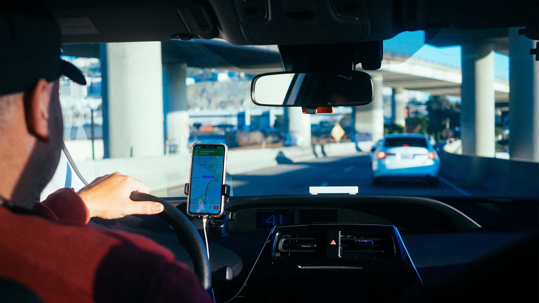 Motorista dirige carro com celular preso ao painel exibindo mapa de navegação, em uma via elevada com tráfego ao fundo