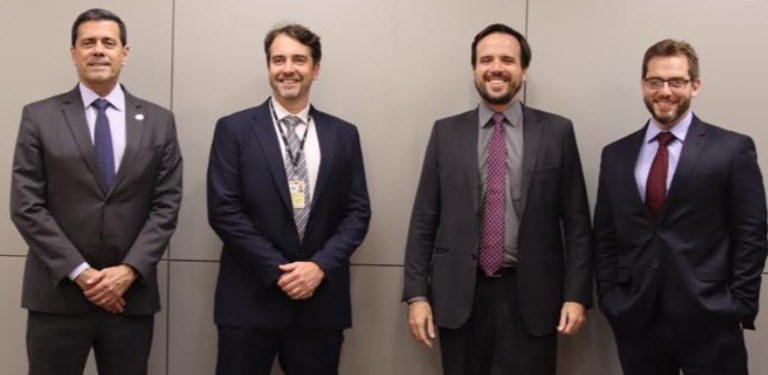 Quatro homens de terno em pé lado a lado diante de parede cinza, posando sorridentes para foto de grupo em evento institucional