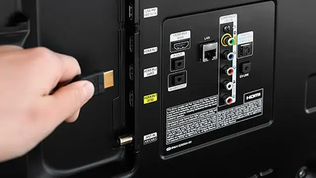 Mão conectando cabo HDMI dourado na entrada HDMI IN ARC amarela de uma TV preta, com painel de portas traseiro visível
