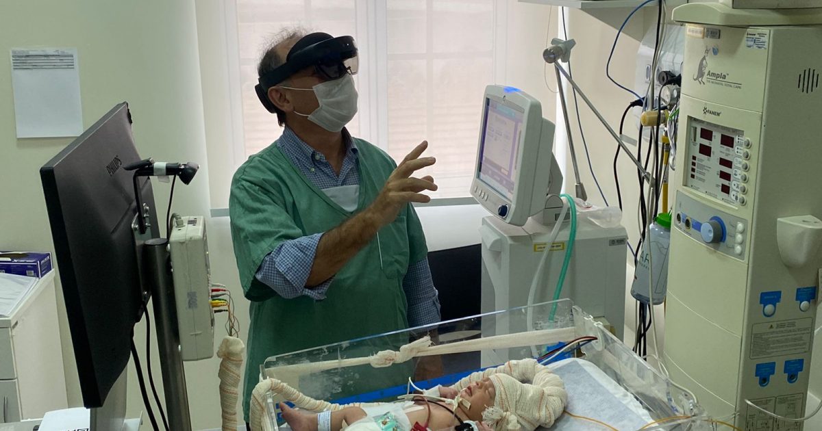 Médico com óculos de realidade mista gesticula sobre uma incubadora de bebê em UTI neonatal, ao lado de monitores e aparelhos hospitalares