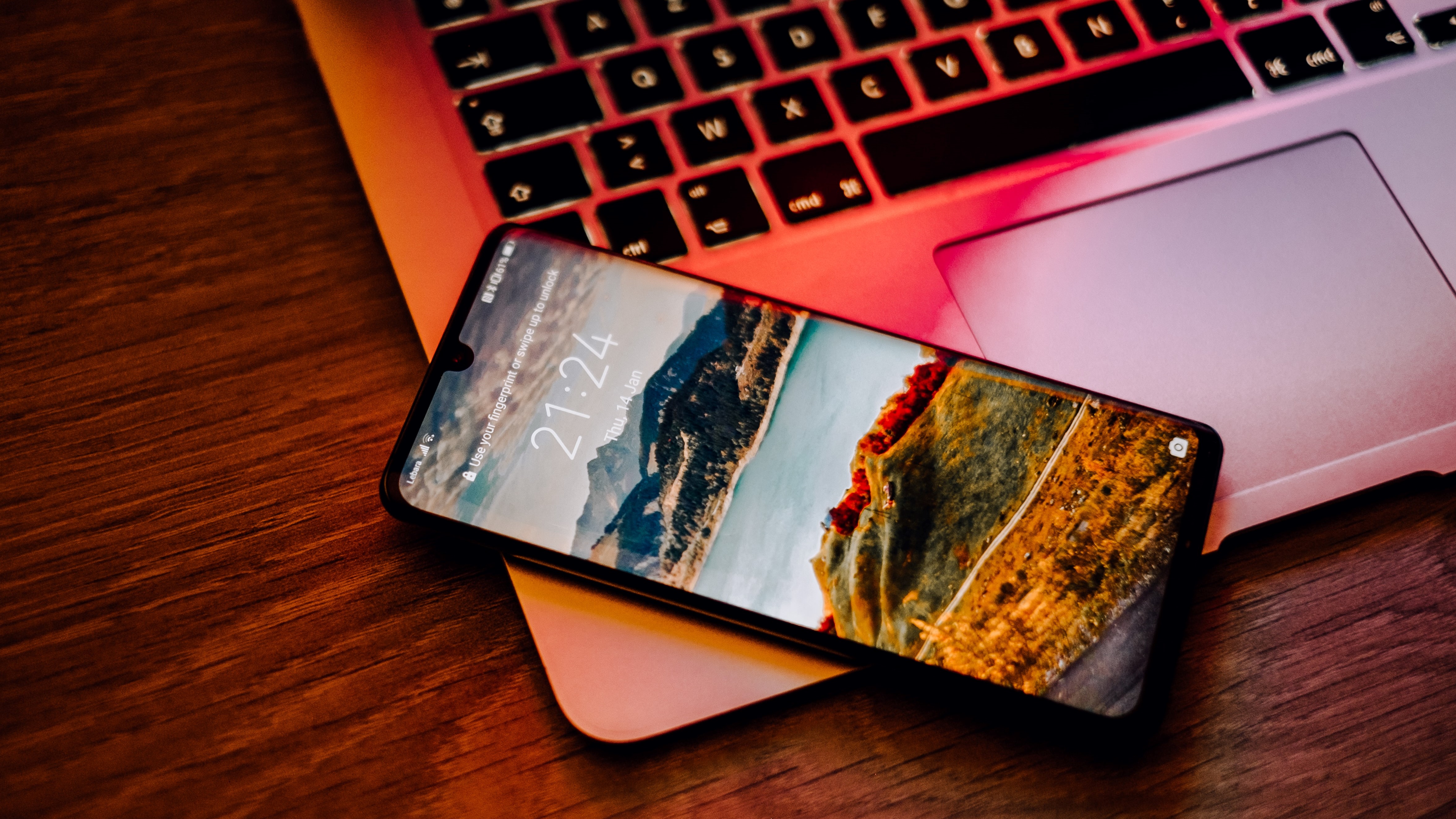 Smartphone com tela de bloqueio exibindo paisagem de montanhas, deitado sobre um notebook com teclado iluminado em tons avermelhados