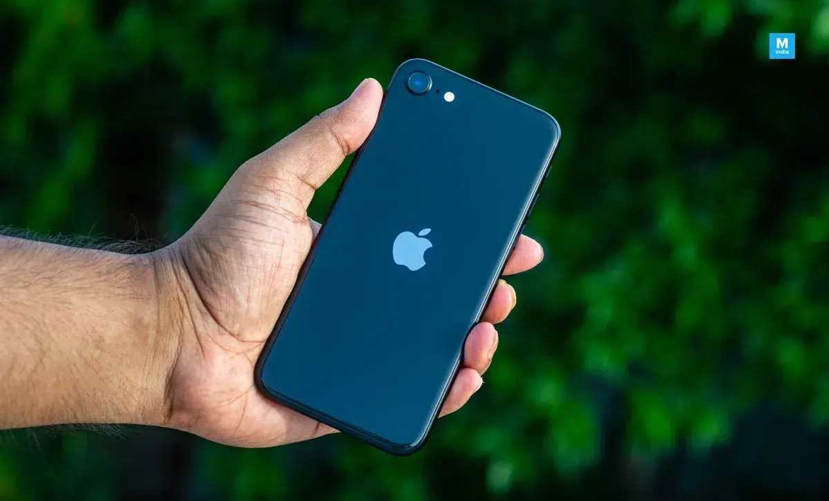 Mão segurando um iPhone SE preto de costas, com logo da Apple e câmera traseira visíveis, diante de fundo verde desfocado