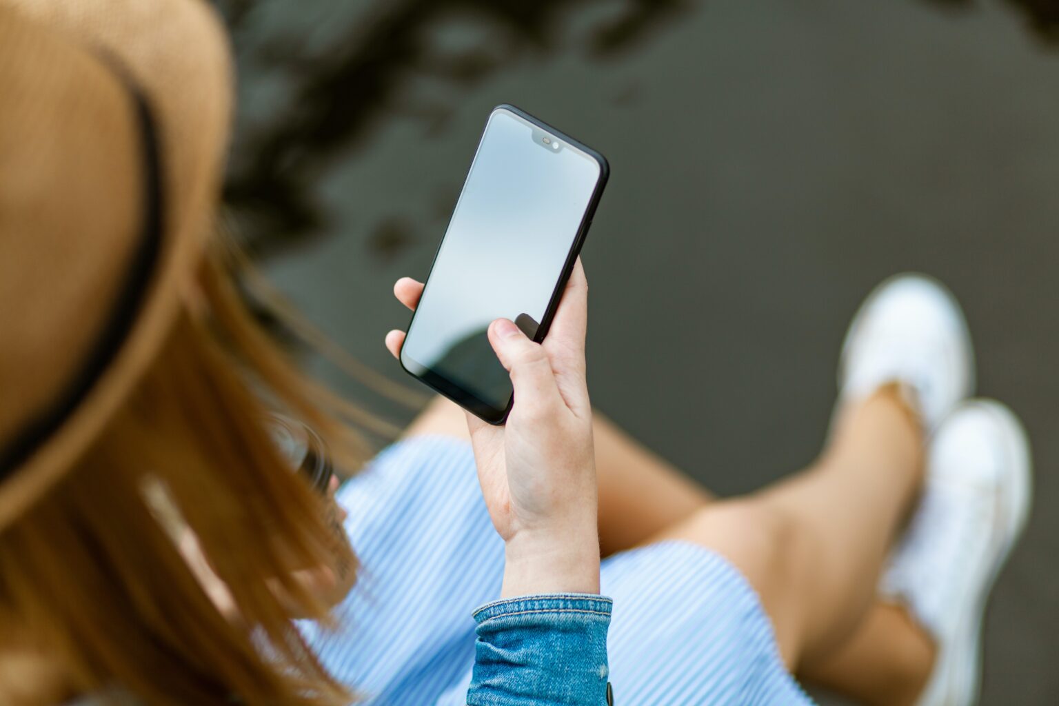 Mulher de chapéu de palha e vestido listrado sentada à beira da água, usando um smartphone com a tela em branco