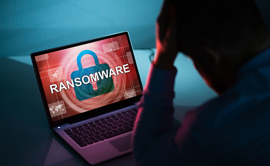 Pessoa de cabeça baixa diante de notebook com a palavra RANSOMWARE e um cadeado sobre fundo vermelho na tela