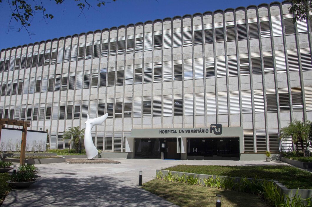 Fachada do Hospital Universitário da USP, prédio de concreto com brises verticais, placa com o nome da instituição e área externa com jardim e escultura branca