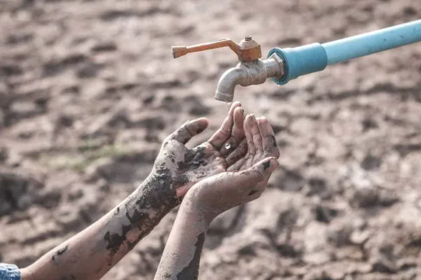 Mãos sujas de terra recebendo água de uma torneira azul em solo rachado e seco, imagem que ilustra a escassez de água